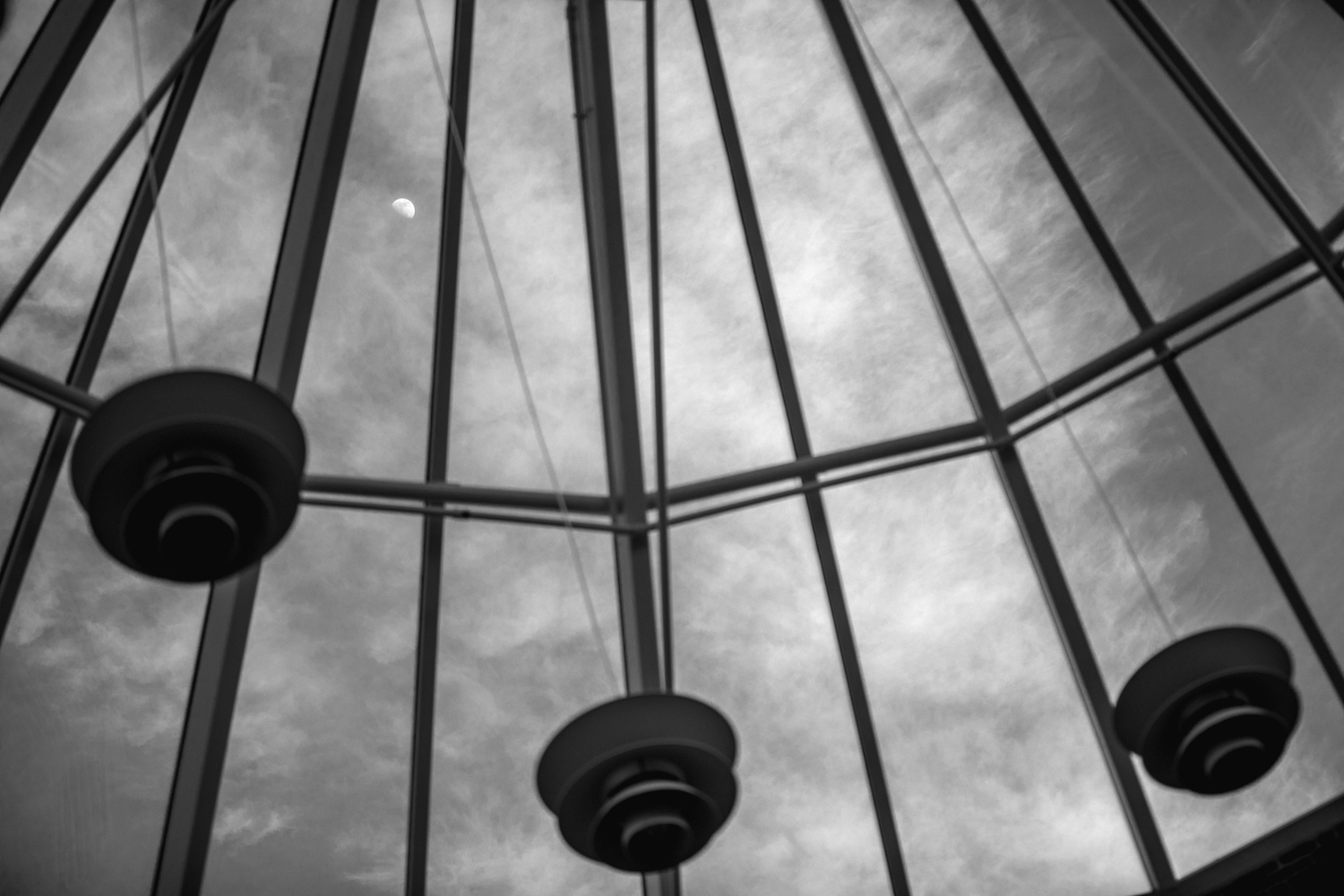 Looking up through a glass dome with leading lines and blue sky, the moon visible up ahead.